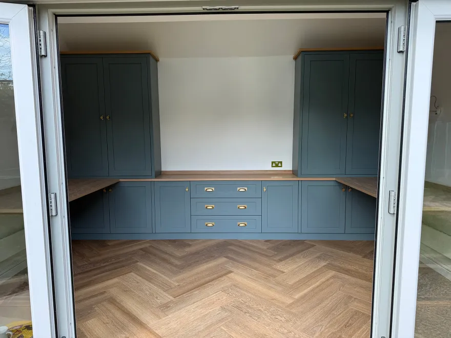 Blue-grey fitted home office seen through open bifold doors, with a herringbone floor