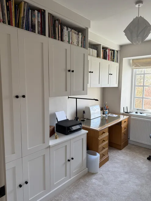 White shaker fitted office cupboards with book shelving above and a desk by the window