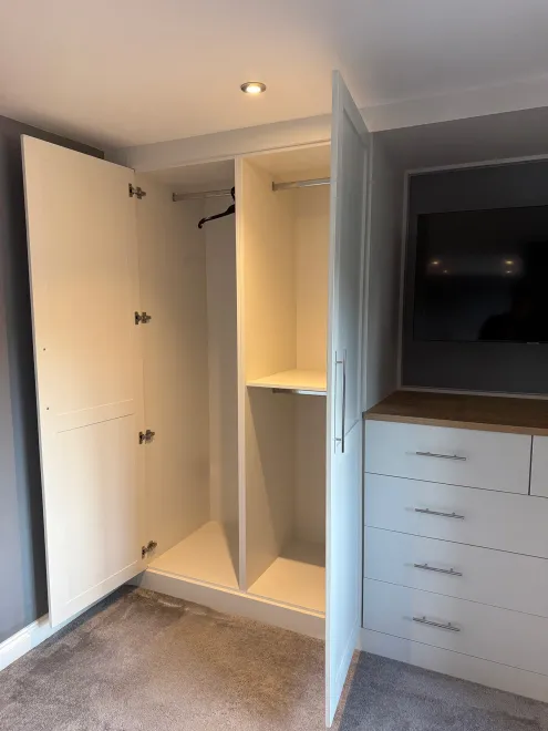 Fitted wardrobe open to show hanging rails, beside a drawer unit and TV recess