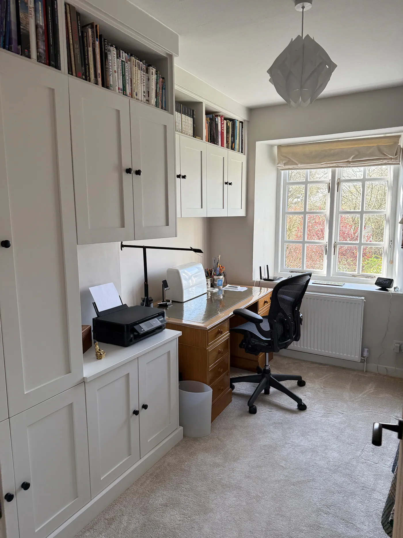 Home office with white fitted cupboards, bookshelves and a desk beneath a window